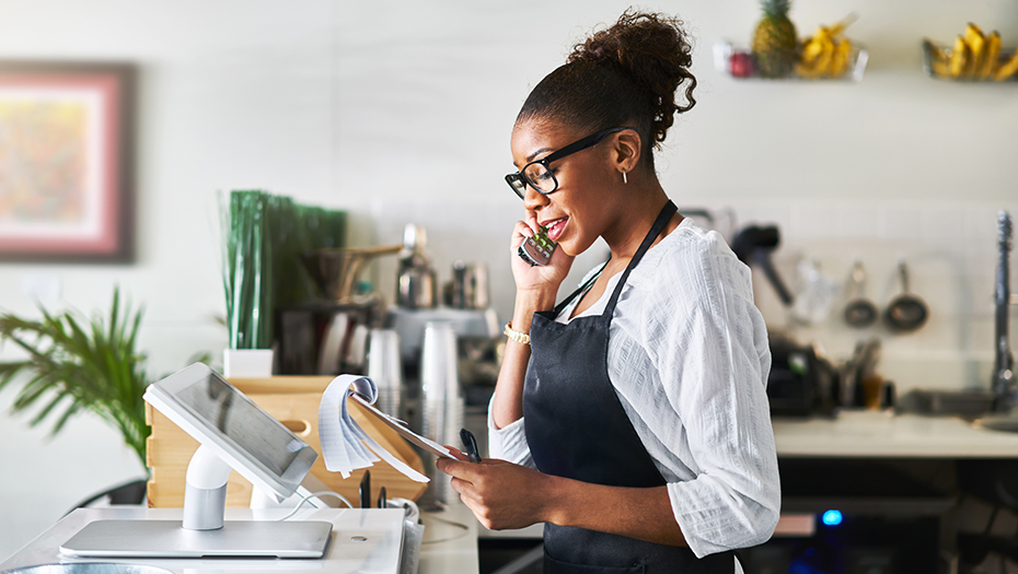 Female small business owner standing behind the cash register, talking on the phone, and reading from a notepad.